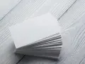 Stos białych wizytówek, idealnych na papier satynowy lub kredowy, leży na drewnianym blacie.