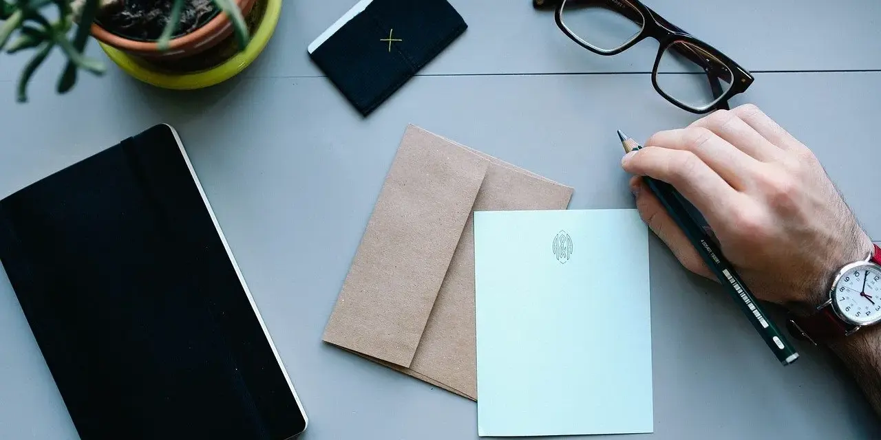 Dłoń z ołówkiem przygotowuje adresowanie koperty. Obok notatnik, okulary i papeteria.