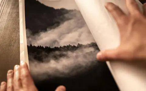 Dłonie otwierają album z fotografią górskiego lasu we mgle, wydrukowaną na papierze barytowym.