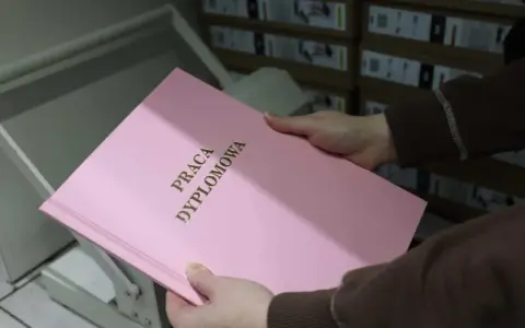 Różowa okładka z napisem "PRACA DYPLOMOWA" w złotych literach, trzymana w dłoniach nad bindownicą.