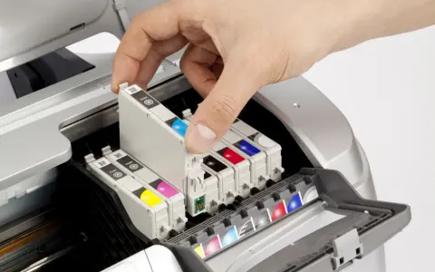 Dłoń wkłada kartridż do drukarki atramentowej. Widać kolorowe tusze i mechanizm budowy drukarki.