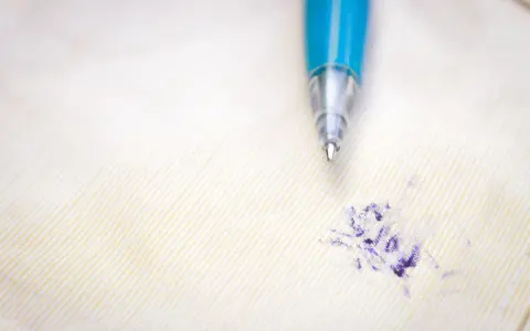 Niebieski długopis nad plamą z atramentu na jasnym materiale. Dowiedz się, jak usunąć pieczątkę z papieru, gdy zdarzy się wypadek.