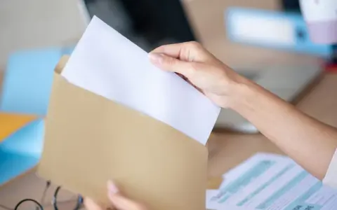 Dłoń wkłada biały papier do brązowej koperty. Wzór adresowania koperty A4 jest gotowy do wypełnienia.