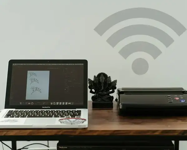 Laptop z projektem graficznym i drukarka obok figurki Ganesha. Symbol Wi-Fi nad nimi sugeruje bezprzewodowe drukowanie.